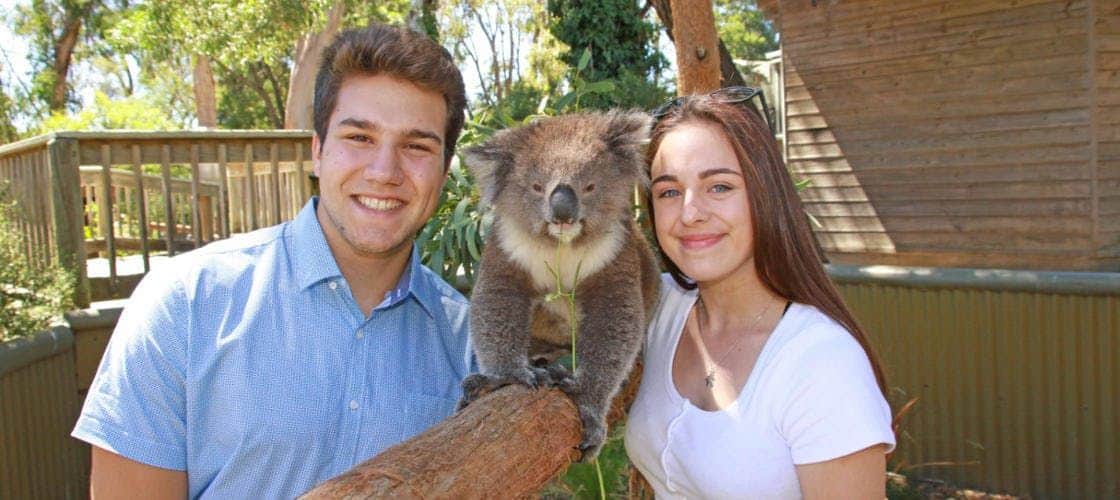 Ballarat Wildlife Park: Entry Ticket Two people smiling while standing next to a koala sitting on a branch in a lush, outdoor setting.