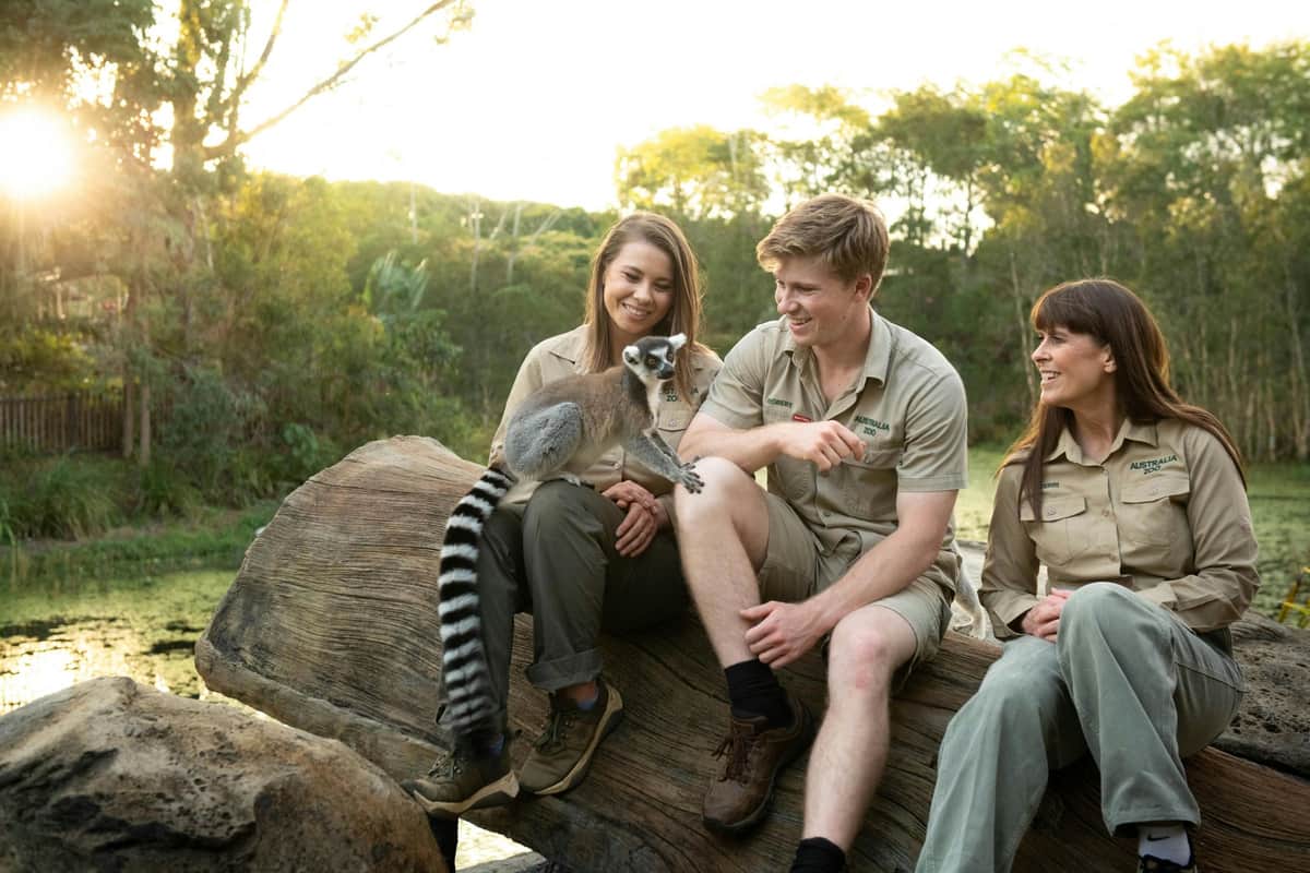 Australia Zoo: Fast Track Entry Ticket + Transfer from Sunshine Coast/Noosa Three people wearing khaki uniforms sit on a log outdoors, smiling at a ring-tailed lemur perched on one person's knee.