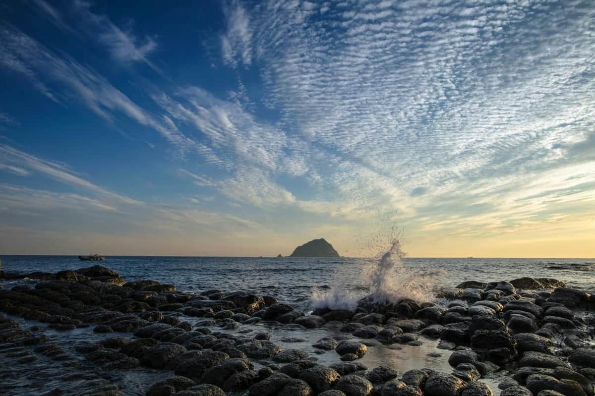 Ondas a rebentar nas rochas arredondadas ao longo da costa, com uma ilha ao longe sob um céu repleto de nuvens dispersas.