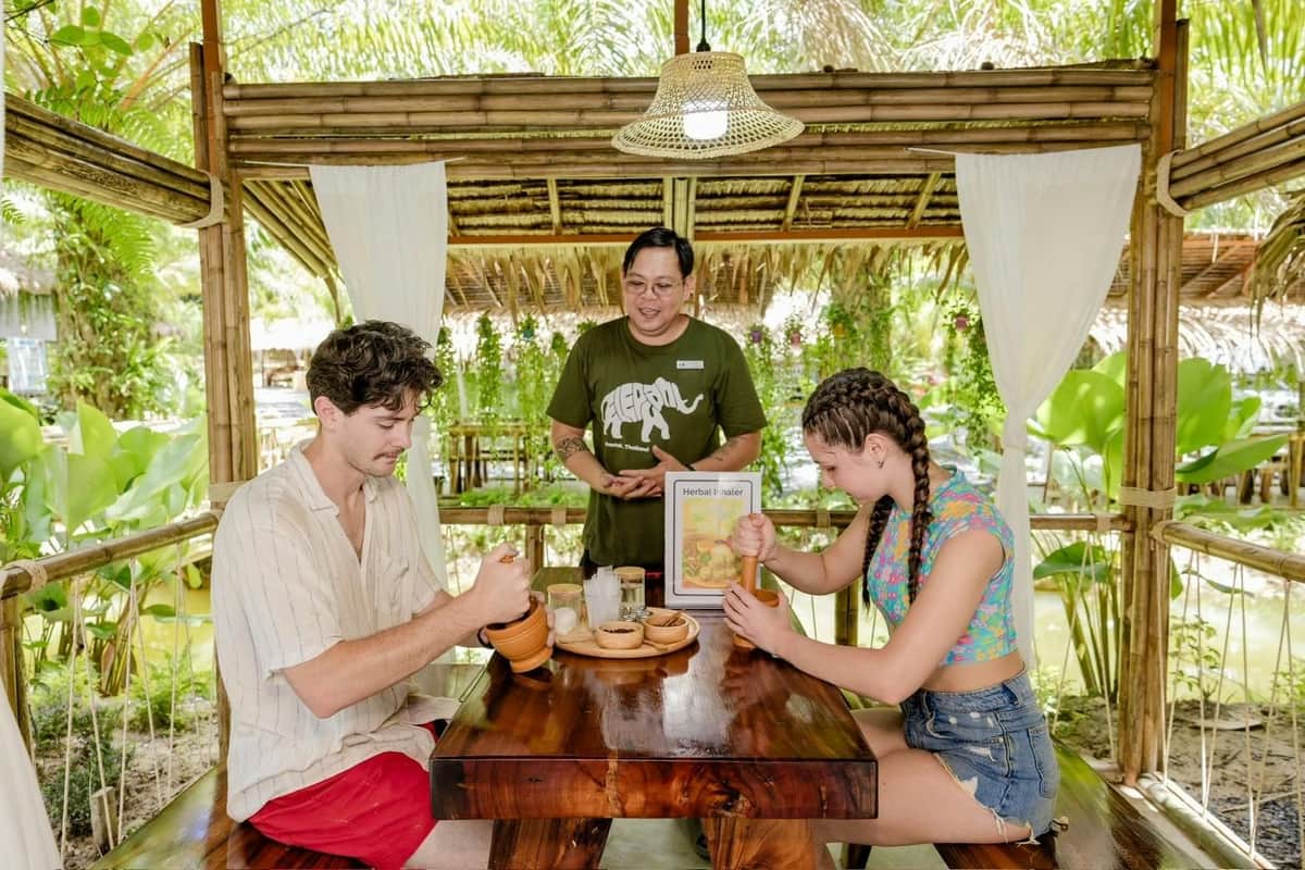 Khaolak Ethical Elephant Sanctuary: Guided Tour + Transfer + Thai Workshops A couple dining at a rustic outdoor restaurant being served by a waiter, surrounded by greenery and wooden decor.
