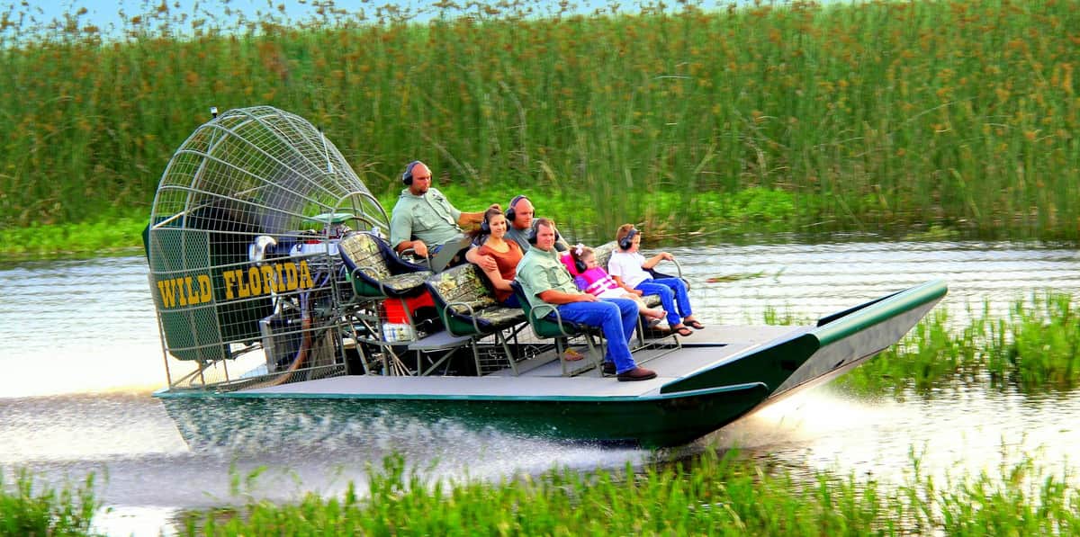 Wild Florida 30-minute Everglades Airboat Tour People wearing headsets riding an airboat labeled "Wild Florida" through tall marshy grass.