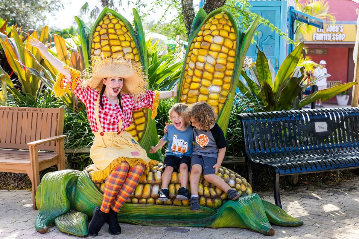 Child in a red hat and overalls smiles next to someone in a manatee costume, with hay bales and a large corn display in the background.