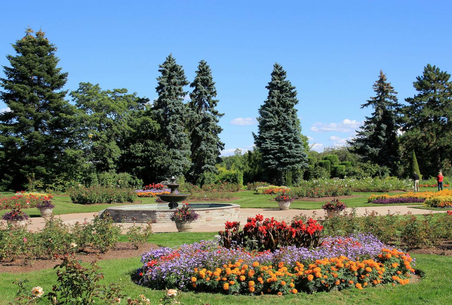 Niagara Falls Butterfly Conservatory Colorful flower beds, a stone fountain, and tall trees in a landscaped park under a clear blue sky, with two people in the distance.