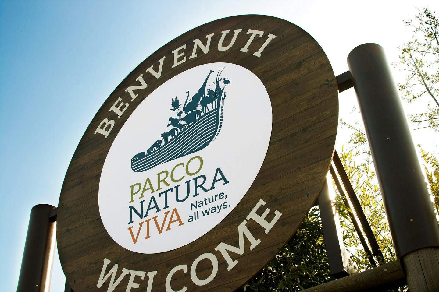 둥근 나무 간판에는 'BENVENUTI' 와 'WELCOME' 이라고 쓰여 있으며, 동물 로고와 'PARCO NATURA VIVA, Nature, all ways' 라는 문구가 새겨져 있습니다.