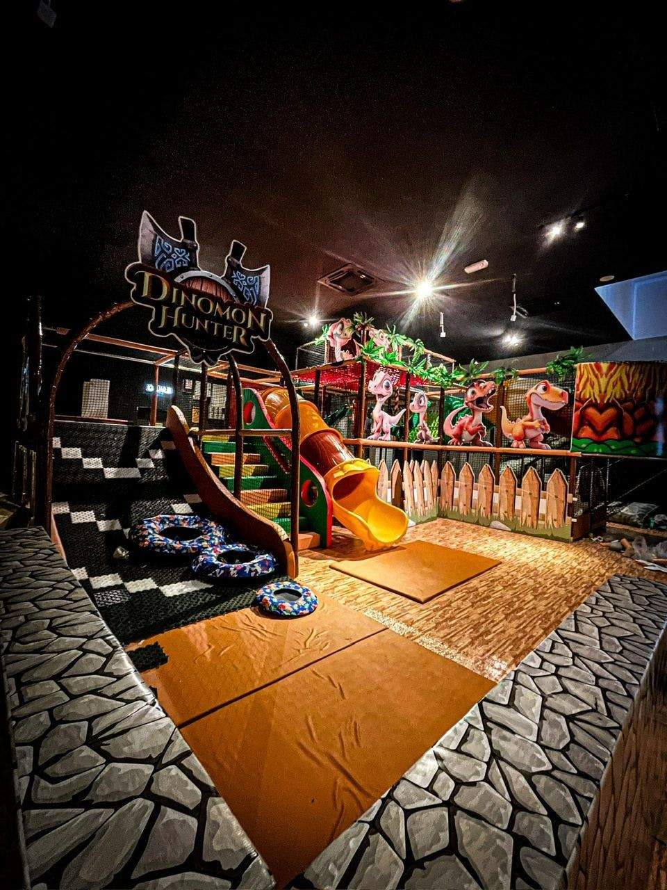 Indoor children's play area with a jungle theme, slides, climbing structures, soft mats, and colorful dinosaur decorations.