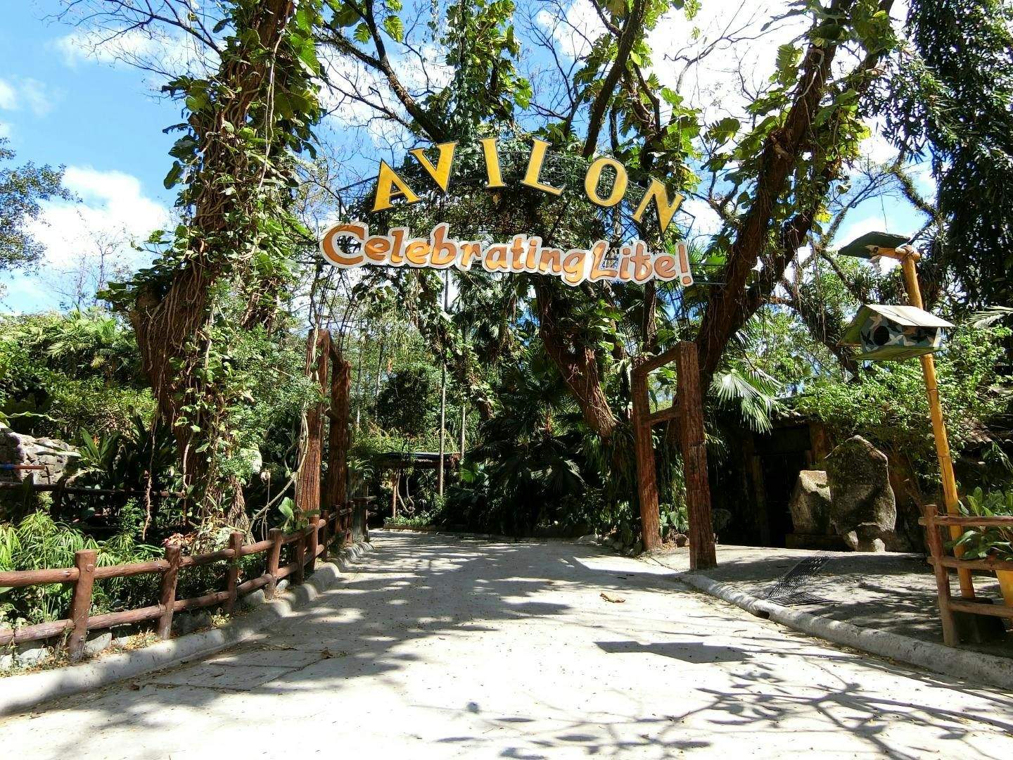 A pathway surrounded by lush greenery with a sign overhead reading 'Avilon Celebrating Life!,' under a sunny, blue sky.