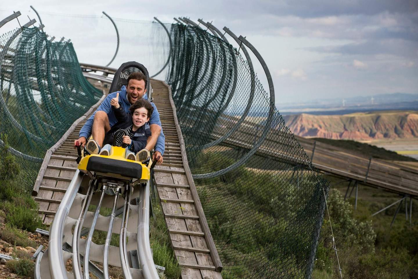 Sendaviva A man and child ride a yellow alpine coaster down a wooden track, surrounded by netting, with mountainous terrain in the background.