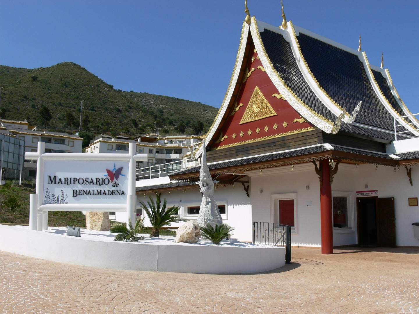 Thai-style building with ornate roof, sign reading "Mariposario de Benalmadena," plants, and mountainous background under a clear blue sky.