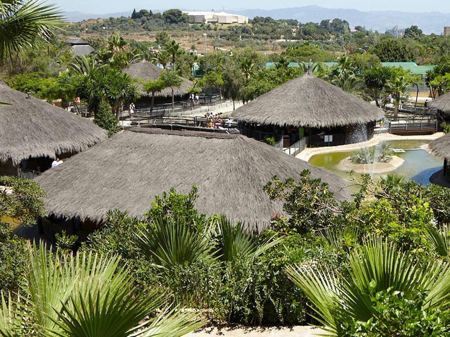 Crocodile Park Torremolinos Thatched-roof huts surrounded by lush greenery and water features in a tropical setting, with people visible in the distance.