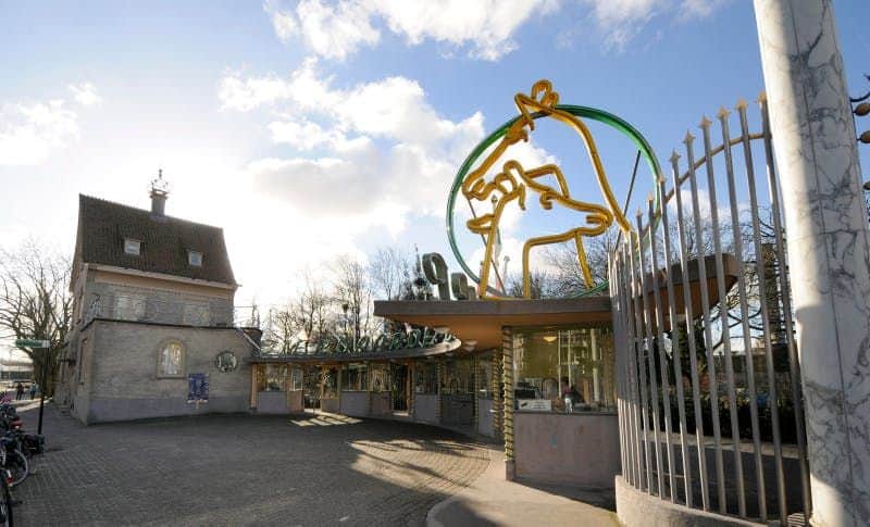 Blijdorp Rotterdam Zoo Entrance gate with decorative sculpture and a ticket booth to the right. A brick building and bicycles are visible to the left.