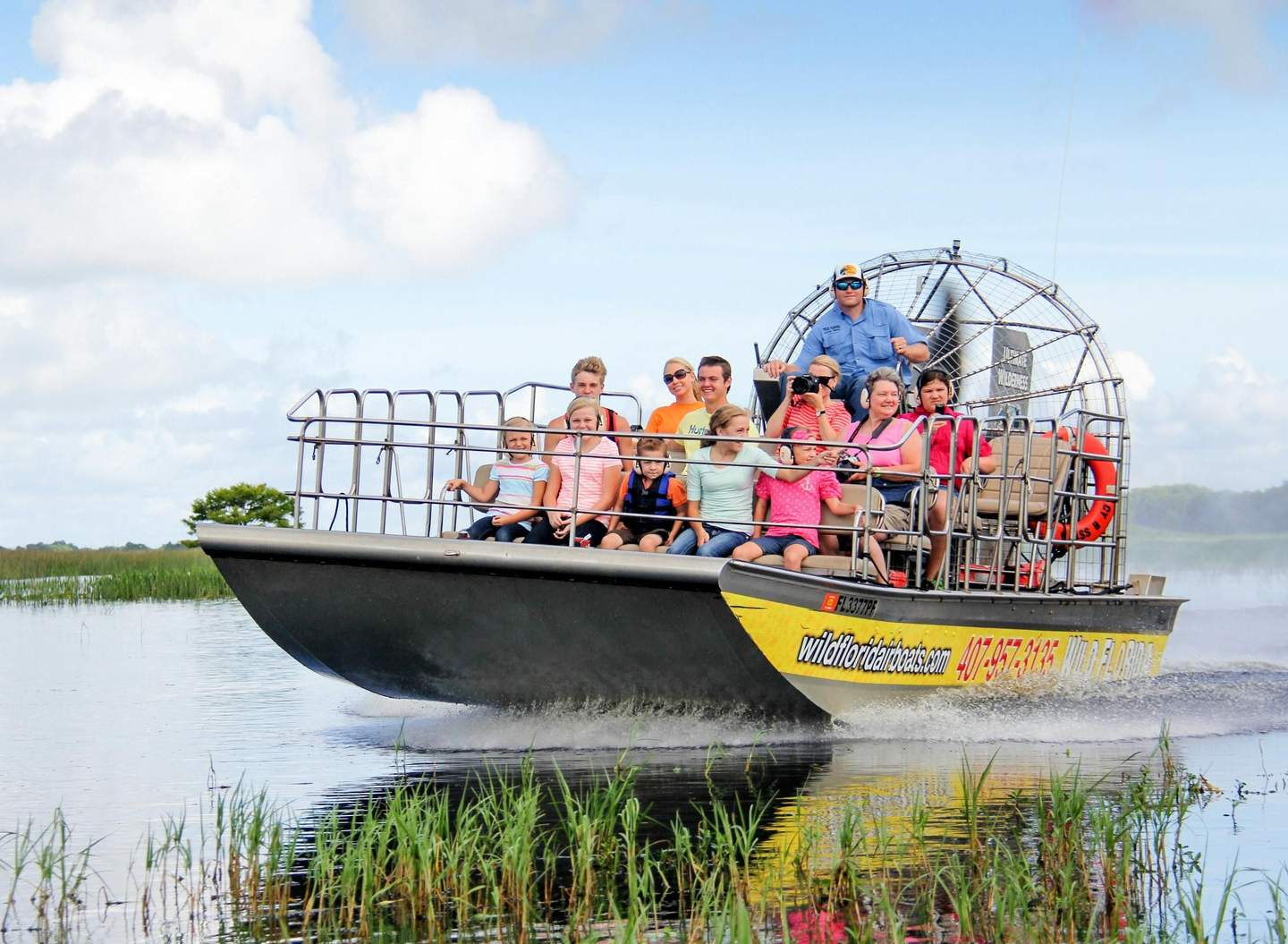 Wild Florida A group of people enjoy an airboat ride through wetlands on a sunny day, with safety railings and a visible propeller in the background.